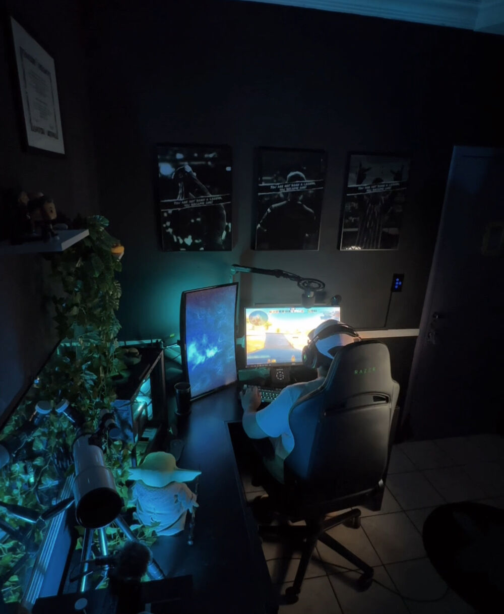 Tonny PK jogando no seu setup: mesa em L com dois monitores, cadeira Razer, microfone em braço articulado, iluminação azul e plantas